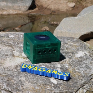 May include: A green Nintendo GameCube console with a black disc drive and four controller ports. The console is sitting on a gray rock with the text "DNAGAMES" in blue and yellow letters.