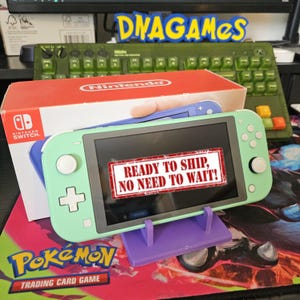 May include: A light green Nintendo Switch Lite handheld gaming console with white buttons and a black screen, positioned in a purple plastic stand in front of its original white and red retail box, featuring the text READY TO SHIP, NO NEED TO WAIT!