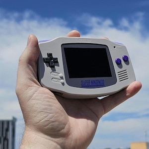 May include: A white and gray handheld gaming console with a black screen and purple buttons. The console has the text "SUPER NINTENDO" printed on the front.