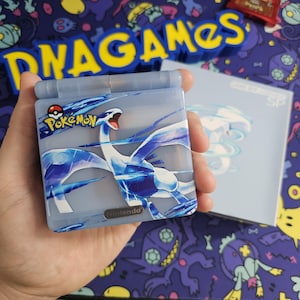 May include: A blue Game Boy Advance SP with a clear plastic shell featuring a Pokemon illustration of a white and blue legendary Pokemon,  "Lugia", with the text "Pokemon" and "Nintendo" on the front.