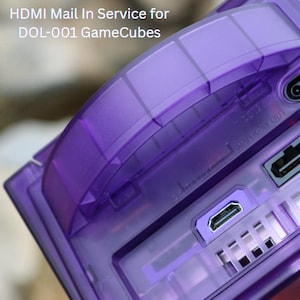 May include: A close-up of a purple GameCube console with the text "HDMI Mail In Service for DOL-001 GameCubes." The console features an HDMI port and other connection ports. The console is made of translucent purple plastic.