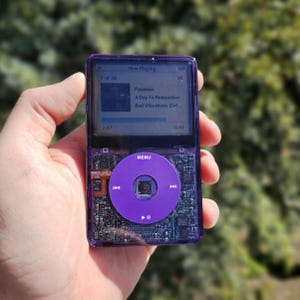 May include: A purple iPod Classic, held in a hand, with a transparent casing revealing internal components. The screen displays the "Now Playing" interface, showing song details for "Paranoia". The click wheel and buttons are purple.