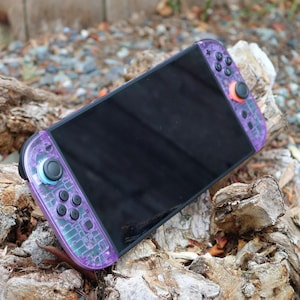 May include: A purple and clear Nintendo Switch Joy-Con controller with black buttons and a black screen. The controller has a circuit board design and orange and blue accents. The device is resting on a piece of wood.