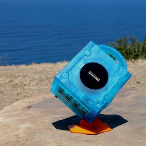 May include: A transparent blue Nintendo GameCube console with a black disc drive cover. The console is propped up on an orange stand against a backdrop of a blue ocean and a grassy hill. The GameCube logo is visible.