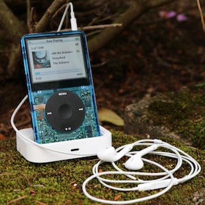 May include: A blue and clear iPod with a white charging dock and white earbuds. The iPod displays the song "The Sickness" by Disturbed. The iPod is resting on a bed of moss.