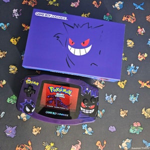 May include: A purple Game Boy Advance game cartridge with a black and white image of a Pokemon character on the front. The cartridge is labeled "Pokemon Ruby Version".