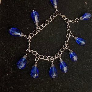 May include: A silver-toned chain bracelet with multiple dangling, faceted, teardrop-shaped, royal blue crystal beads. The bracelet is displayed on a black surface, showcasing the contrast between the silver and blue.