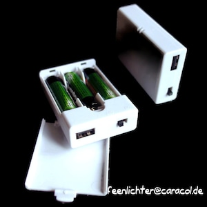 Può includere: Un pacco batterie in plastica bianca con tre batterie AA inserite. Il pacco ha una porta USB e un pulsante di accensione.