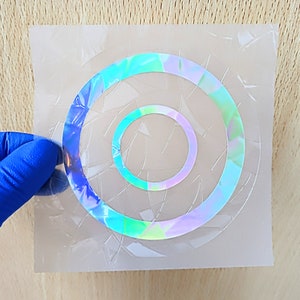 Pode incluir: Uma folha de plástico transparente com um adesivo holográfico iridescente de dois círculos concêntricos. O adesivo tem um efeito de vidro rachado.