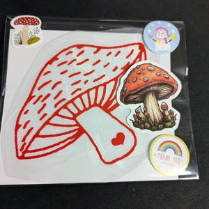 May include: A package of stickers featuring mushroom designs. The main sticker is a large red and white mushroom outline. Other stickers include a detailed orange mushroom, a snowman, and a rainbow with the words "Thank You".