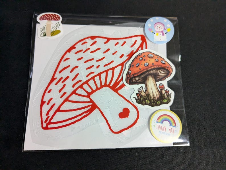 May include: A package of stickers featuring mushroom designs. The main sticker is a large red and white mushroom outline. Other stickers include a detailed orange mushroom, a snowman, and a rainbow with the words "Thank You".