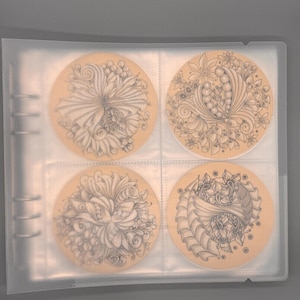 May include: Four round coasters with intricate black and white floral designs are displayed in a clear plastic holder. The coasters have a light tan background and are arranged in a two-by-two grid.