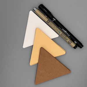 May include: An overhead shot shows a collection of triangular paper pieces in white, tan, and brown, alongside two black pens. The pens are labeled "MICRON PN" and "uni PIN FINE LINE 0.05". The items are arranged on a gray surface.