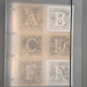 May include: A clear plastic binder with a black elastic closure, holding six square cards. The cards feature ornate letter designs, including A, B, C, E, and F, with floral and decorative elements. The binder is on a gray background.