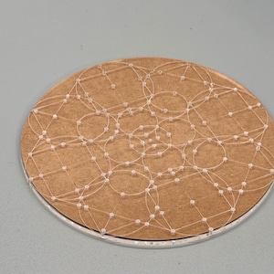 May include: A round, brown coaster with a network of white thread and small, clear beads. The thread forms interconnected circles across the surface. The coaster has a silver-colored edge and sits against a gray background.