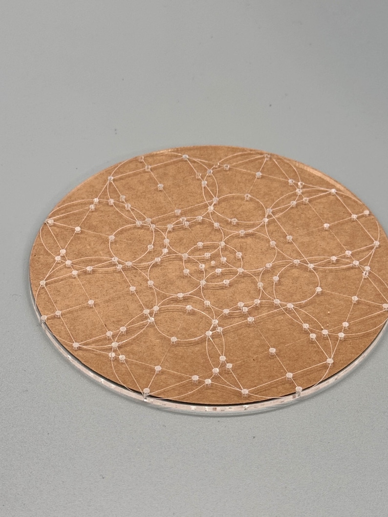 May include: A round, brown coaster with a network of white thread and small, clear beads. The thread forms interconnected circles across the surface. The coaster has a silver-colored edge and sits against a gray background.
