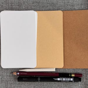 May include: Three blank notepads, one white, one beige, and one brown, are stacked on top of each other. A white pencil, a red pencil, and a black marker are lying on top of the notepads.