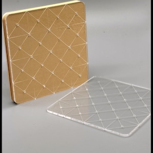 May include: A square, tan-colored notebook with a clear, grid-like design made of string and small beads. A separate, clear acrylic sheet with the same grid pattern is also present. The notebook is open, revealing the pages.
