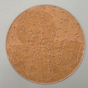 May include: A round, brown wooden coaster with a geometric design. The design features interconnected circles, squares, and lines, creating a complex pattern. The coaster is made of a light brown wood material.