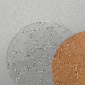May include: Two clear, circular stencils with geometric designs. One stencil is transparent, and the other is a light brown color. The stencils are placed on a gray surface.