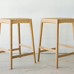 May include: Two wooden stools with woven seats. The stools have a simple, modern design and are made of light-colored wood.