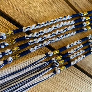 Peut inclure: Gros plan sur plusieurs pompons faits à la main sur le thème nautique. Chaque pompon est composé d'un cordon torsadé blanc et bleu marine, orné de perles métalliques dorées. Les pompons sont disposés sur une surface en bois.