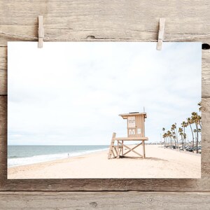 May include: A photographic print featuring a light tan wooden lifeguard tower on a sandy beach with the ocean in the background. The tower has a white letter M on the front. Tall palm trees line the distant shore under a bright, overcast sky.