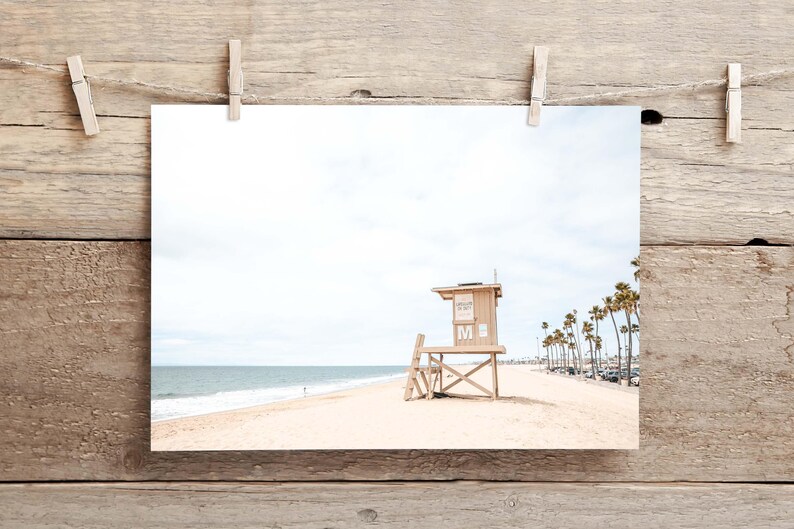 May include: A photographic print featuring a light tan wooden lifeguard tower on a sandy beach with the ocean in the background. The tower has a white letter M on the front. Tall palm trees line the distant shore under a bright, overcast sky.