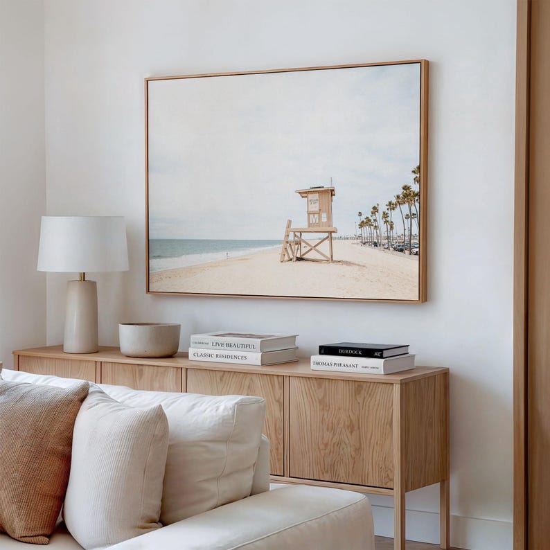 May include: A rectangular wall art print in a light wood frame depicting a beach scene with a wooden lifeguard tower, sandy shore, ocean, and palm trees under a pale sky. The artwork is displayed above a light wood console table with books.