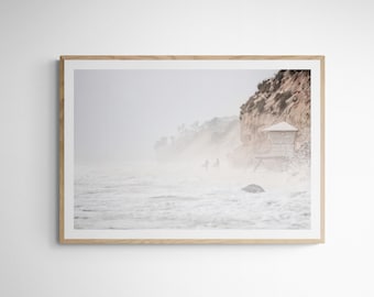 May include: A foggy beach scene with a small building on a cliff. The water is white with foam and there are two figures walking in the distance.