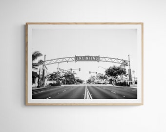 May include: Black and white photograph of a street sign that reads "ENCINITAS" over a road with cars and buildings on either side.