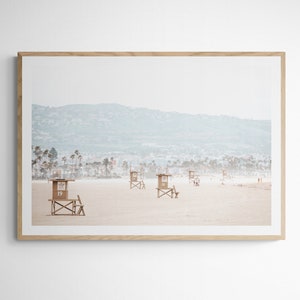 May include: A photograph of a beach with lifeguard stands in the foreground and a hazy mountain range in the background. The beach is sandy and there are a few people walking on it.