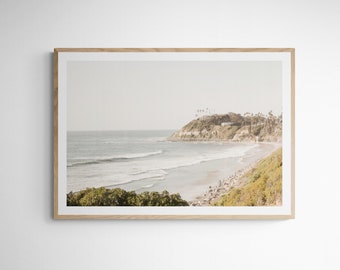 May include: A framed photograph of a coastal scene with a sandy beach, rolling waves, and a green hillside with trees and houses in the distance.