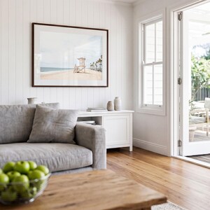 May include: A framed landscape photograph of a beach with a lifeguard tower and palm trees, displayed on a white vertical paneled wall above a white media console in a living room with a gray sofa and a wooden coffee table holding a bowl of green apples.
