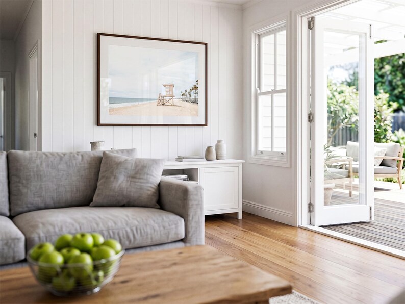 May include: A framed landscape photograph of a beach with a lifeguard tower and palm trees, displayed on a white vertical paneled wall above a white media console in a living room with a gray sofa and a wooden coffee table holding a bowl of green apples.