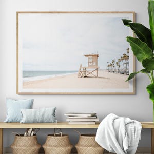 May include: A framed horizontal wall art print depicting a sandy beach scene with a wooden lifeguard tower, calm ocean waves, and a row of palm trees under a pale sky. The print is displayed in a light wood frame on a white wall above a wooden bench.
