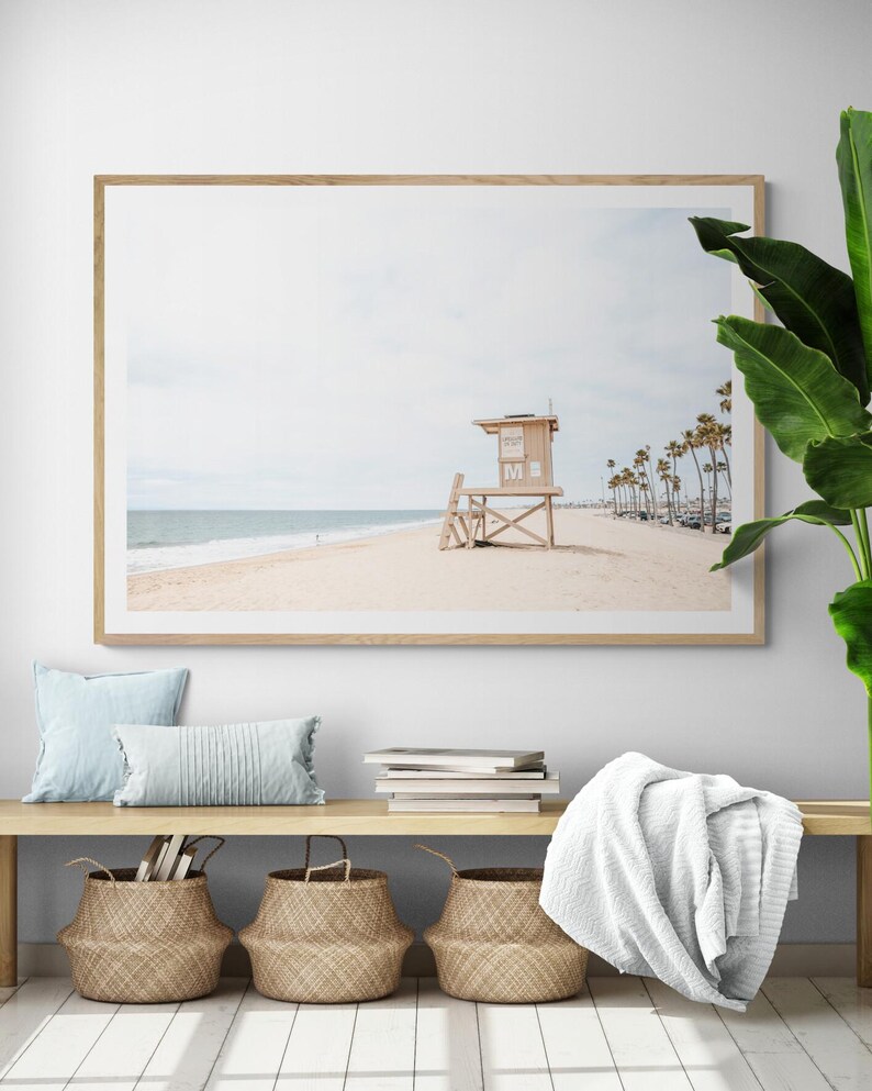 May include: A framed horizontal wall art print depicting a sandy beach scene with a wooden lifeguard tower, calm ocean waves, and a row of palm trees under a pale sky. The print is displayed in a light wood frame on a white wall above a wooden bench.