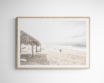 Może przedstawiać: Zdj?cie w ramie przedstawiaj?ce pla?? z konstrukcj? dachu ze strzechy na pierwszym planie i surferem id?cym po pla??y w oddali. Niebo jest pochmurne, a ocean ma jasnoniebieski kolor.