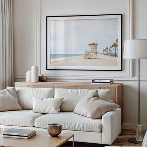 May include: A large framed horizontal wall art print depicting a sandy beach with a wooden lifeguard tower and palm trees under a pale sky. The artwork is displayed above a light wood console table and a cream-colored sofa with matching throw pillows.