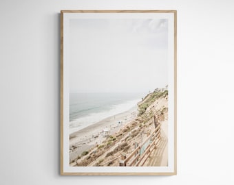 May include: A photograph of a beach scene with a wooden fence and a path leading down to the beach. The beach is sandy and the water is blue. The sky is cloudy and the sun is shining.