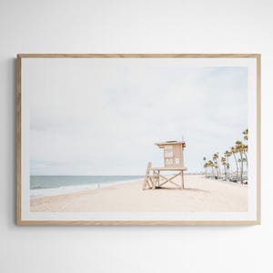 May include: A framed landscape photograph featuring a light-colored wooden lifeguard tower on a sandy beach. The scene includes the ocean, a pale sky, and a row of palm trees in the distance. The frame is made of light-toned wood with a white mat.