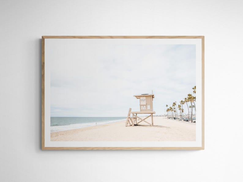 May include: A framed landscape photograph featuring a light-colored wooden lifeguard tower on a sandy beach. The scene includes the ocean, a pale sky, and a row of palm trees in the distance. The frame is made of light-toned wood with a white mat.