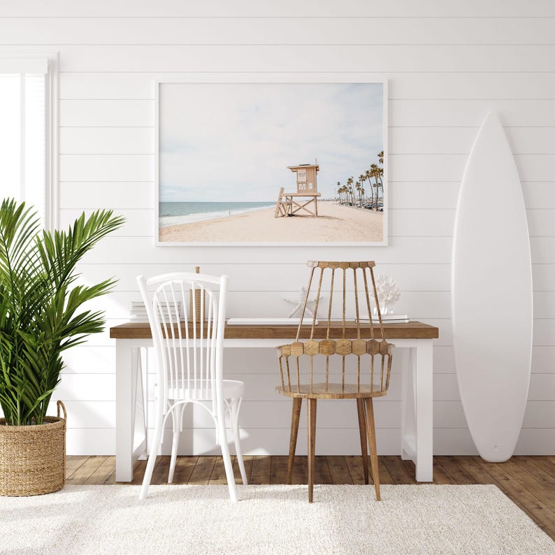 May include: A rectangular framed wall art print depicting a beach scene with a lifeguard tower and palm trees, positioned above a wooden desk with a white spindle chair, a brown wooden chair, a green potted plant, and a white surfboard.