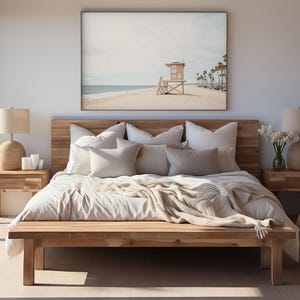 May include: A bedroom featuring a wooden bed frame with white bedding and multiple pillows, flanked by two wooden nightstands with beige lamps. Above the bed hangs a large framed print of a beach scene with a lifeguard tower and palm trees.