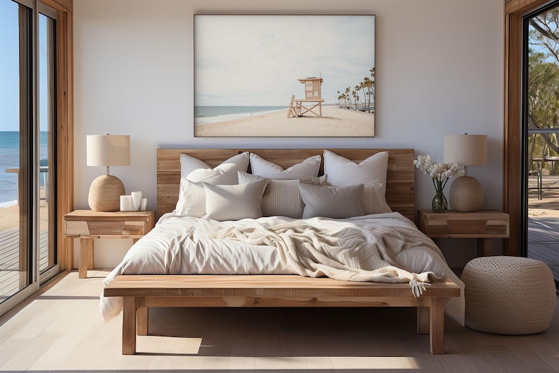 May include: A bedroom featuring a wooden bed frame with white bedding and multiple pillows, flanked by two wooden nightstands with beige lamps. Above the bed hangs a large framed print of a beach scene with a lifeguard tower and palm trees.