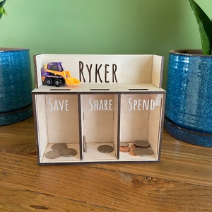 May include: Wooden coin bank with three compartments labeled "Save", "Share", and "Spend". The bank has a small toy construction vehicle on top and the name "Ryker" written on the front.