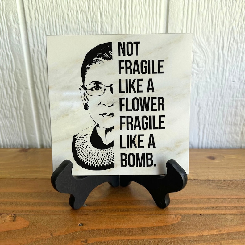 May include: A white ceramic tile with a black and white image of Ruth Bader Ginsburg and the text "Not fragile like a flower fragile like a bomb."