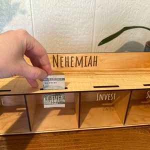 May include: A wooden money organizer with four labeled compartments: SAVE, GIVE, INVEST, and SPEND. The name "NEHEMIAH" is engraved on the top. A hand is placing a folded dollar bill into the "GIVE" slot. The organizer is made of light-colored wood.