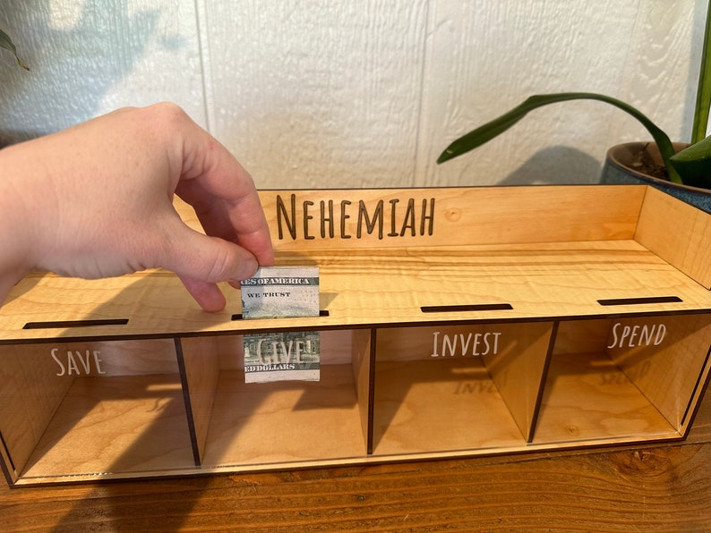 May include: A wooden money organizer with four labeled compartments: SAVE, GIVE, INVEST, and SPEND. The name "NEHEMIAH" is engraved on the top. A hand is placing a folded dollar bill into the "GIVE" slot. The organizer is made of light-colored wood.