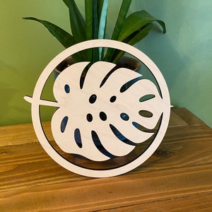 May include: A wooden circle with a cutout of a monstera leaf. The leaf is white and the circle is brown.